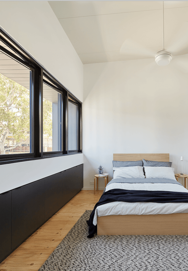 A bright, minimalist bedroom with a low timber bed positioned beside a long bank of black-framed windows. The room has white walls and ceiling, warm timber flooring, dark built-in cabinetry beneath the windows, a textured neutral rug, simple bedside tables, layered white and grey bedding, and a white ceiling fan. Strong natural light enters through the expansive glazing.