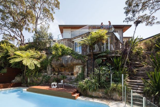 A contemporary multi-level hillside residence viewed from a landscaped pool area. The home features broad roof overhangs, extensive glazing, glass balustrades, timber decking, and layered outdoor terraces integrated into a rocky, planted garden. Stone steps rise through dense vegetation toward the house and rooftop terrace, while a swimming pool occupies the foreground.