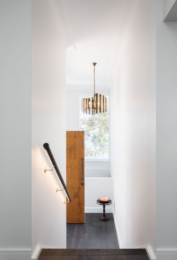 A bright, narrow residential stair landing or entry passage with white walls and ornate ceiling detailing. Dark timber flooring leads toward a tall natural timber door or screen, with a large window beyond. A sculptural black-and-brass handrail runs along the left wall, while a small round black side table and a suspended bronze-toned linear pendant add refined decorative accents.