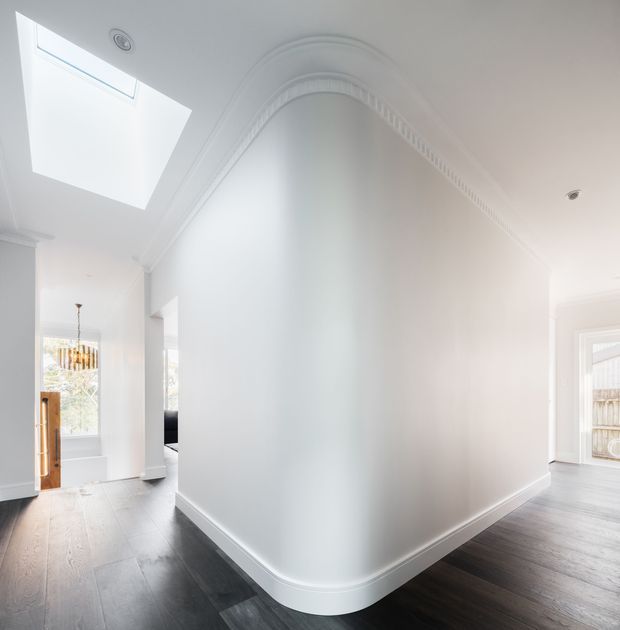 Bright white interior hallway or entry passage with a dramatic curved wall and ornate decorative cornice detailing. Dark timber flooring contrasts with the white walls and trim. A large skylight brings in natural light, with glimpses of a front door, pendant light, and adjacent rooms visible at the sides.