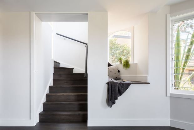 Bright, minimalist interior view of a stairway beside a built-in window seat. The staircase has dark timber treads and a simple dark handrail, contrasting with white walls, white trim, and pale flooring. A large window and arched opening bring in natural light, while the window seat includes neutral cushions and a dark throw.