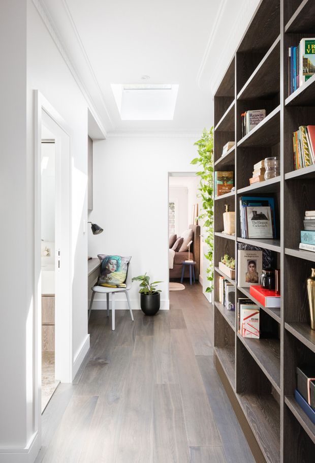 Bright, narrow residential hallway with white walls, decorative ceiling cornices, a large skylight, dark full-height built-in bookcases on the right, and grey-brown timber-look flooring. A small seating nook, potted plants, and an open doorway to a living area are visible beyond. The space has a clean, refined contemporary interior with strong contrast between the white envelope and dark joinery.
