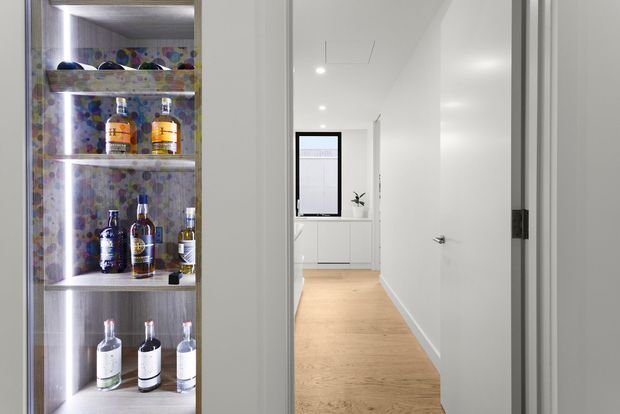 View through a doorway into a bright, minimalist hallway or passage with pale timber flooring, white walls, recessed ceiling lights, and built-in white cabinetry at the far end. A tall open built-in display niche containing bottles occupies the left side, adding a colourful patterned backdrop. A small window and simple decor are visible in the background.