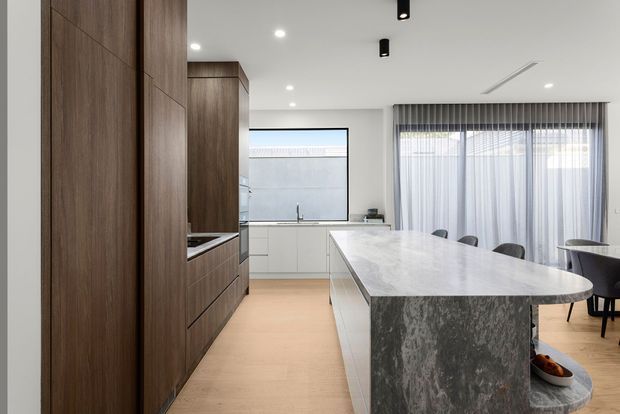 Bright contemporary open-plan kitchen and dining area with a large waterfall-edge stone island, dark timber kitchen cabinetry, white cabinetry, light timber flooring, and a long dining table beside full-height sheer curtains. Recessed and track-style lighting create a clean, refined interior.