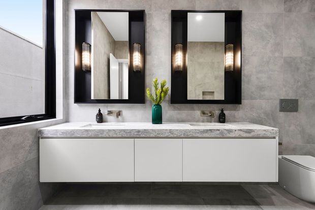 Contemporary bathroom featuring a long floating white vanity with two integrated basins, a grey stone-look benchtop, two black-framed mirrors, wall-mounted sconces, grey tiled walls and floor, and a partially visible toilet. A large window provides natural light, while a small green vase adds colour.
