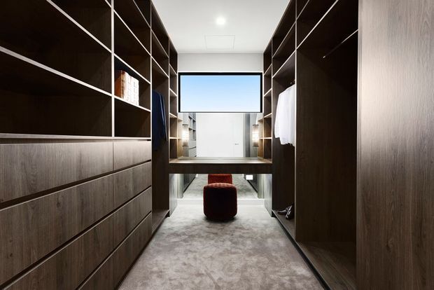 A walk-in wardrobe with full-height dark timber cabinetry, open shelving, drawers, hanging rails, and a central dressing desk beneath a wall-mounted screen or mirror. A plush red ottoman sits at the far end, while soft grey carpet covers the floor. The space is contemporary, streamlined, and carefully integrated with built-in storage.