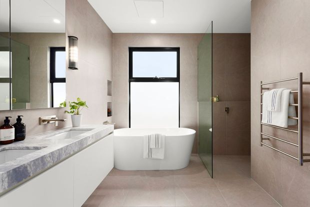 Contemporary bathroom with a freestanding white bathtub positioned beneath a large black-framed window, a floating double vanity with pale stone benchtop, frameless glass shower enclosure, heated towel rail, and large-format warm beige wall and floor tiles. A mirror extends above the vanity, and the room is brightly lit with a clean, refined finish.
