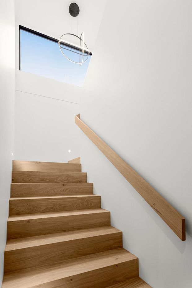 Minimalist interior view of a straight timber staircase ascending toward a bright landing. The stairs and wall-mounted handrail are finished in warm natural wood, set against smooth white walls. A large skylight brings in daylight, and a contemporary circular pendant light hangs overhead.