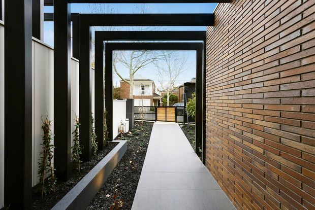 A narrow contemporary covered walkway runs between a dark-framed building facade on the left and a warm reddish-brown exposed brick wall on the right. Repeated black rectangular portal frames create a strong rhythm overhead, with minimalist raised planter beds and dark gravel beside a smooth pale concrete path leading toward a gated courtyard and neighbouring residences.
