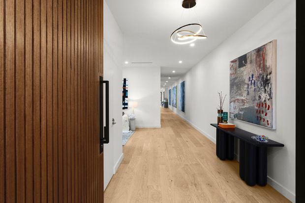 A contemporary residential hallway viewed from an open timber slatted entry door. The long corridor has pale timber flooring, white walls and ceiling, recessed downlights, sculptural pendant lighting, and a dark console table beneath a large abstract artwork. Additional artwork and furnishings extend down the corridor, creating a refined gallery-like entry sequence.