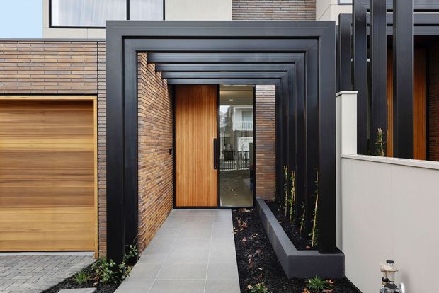 Contemporary residential entry viewed from the front, featuring a deep covered passage framed by repeated dark metal portals, warm timber and glazed entry door, exposed brick walls, large grey floor tiles, and integrated raised planter beds. A timber garage door and sections of the modern facade are visible at the sides.