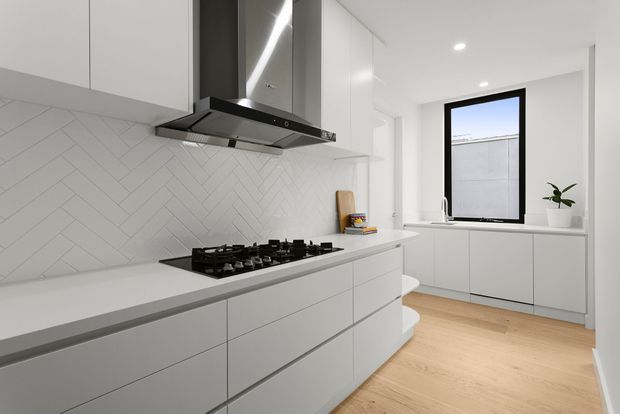 Bright contemporary white kitchen with streamlined handleless cabinetry, a large island with gas cooktop, stainless steel rangehood, and a white herringbone tile splashback. Light timber flooring and a black-framed window add contrast, while integrated cabinetry continues beneath the window.