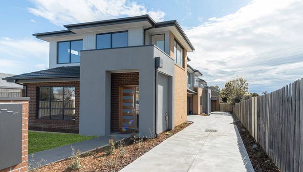 Exterior view of a modern multi-storey townhouse-style residence, featuring a flat/low-pitched roofline, rendered grey facade, exposed brick walls, large dark-framed windows, and a concrete driveway running alongside a timber boundary fence.