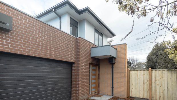 Exterior view of a contemporary multi-level townhouse or residential dwelling. The facade combines red brick and pale rendered upper walls with a dark grey sectional garage door, projecting balcony, timber-and-glass entry door, and a light timber boundary fence. A sharply angled roofline and clean geometric forms define the design.