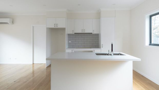 Bright, compact open-plan kitchen with white shaker-style cabinetry, a light stone-look island benchtop, grey subway-tile splashback, integrated sink and dark tapware. Light timber flooring continues into the adjacent open living area, with white walls, recessed lighting and a window providing natural light.