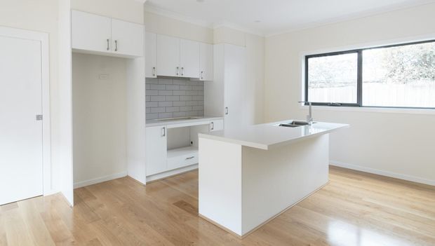 Bright, compact contemporary kitchen with white shaker-style cabinetry, a central island with integrated sink, grey subway-tile splashback, pale timber flooring, and a large black-framed window. The room is minimally furnished and receives abundant natural light.