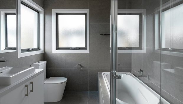 A contemporary bathroom with large-format dark grey wall and floor tiles, white-framed frosted windows, a white vanity with double undermount basins and simple metal pulls, a toilet, and a freestanding-style white bathtub enclosed by a frameless glass shower screen. The space is clean, bright, and practical, with reflective glazing creating additional views of the room.