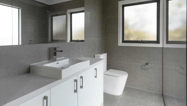 Contemporary bathroom with a white floating vanity, integrated rectangular basin, wall-mounted chrome tapware, toilet, and large-format grey tiled walls and floor. Frosted and translucent windows provide soft natural light, while a large mirror reflects the vanity and room.