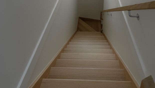 A narrow interior stairway viewed looking upward, with pale timber treads and matching timber handrails mounted to white walls. The stair rises to a small landing and turns at the top, creating a clean, understated composition.