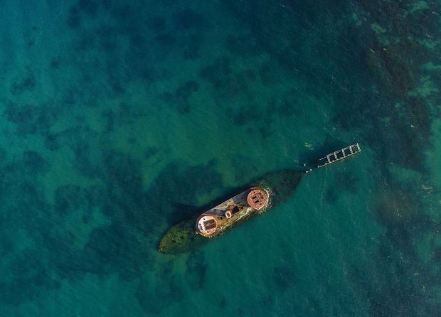 Aerial view looking down at a small, weathered vessel or floating structure surrounded by clear turquoise and deep teal water. A narrow gangway or dock extends from the structure toward the upper right, with underwater seabed and reef-like textures visible through the water. No architectural interior or building envelope is clearly shown.