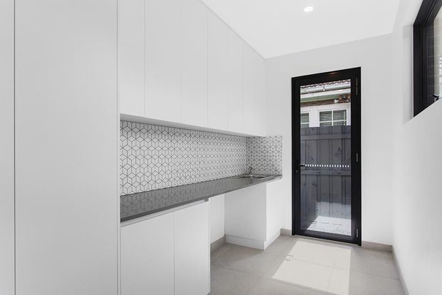 A bright, compact contemporary kitchen or kitchenette with full-height white cabinetry, a slim grey benchtop, and a geometric patterned tiled splashback. A black-framed glazed door and window provide natural light, while large pale floor tiles create a clean, minimal finish.