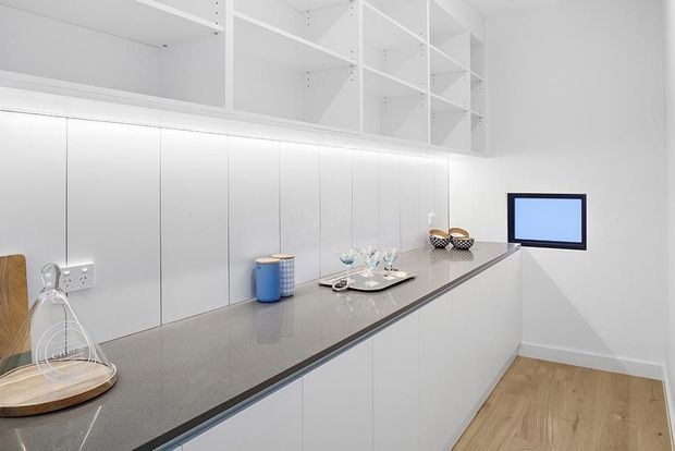 A bright, minimalist kitchen with a long grey benchtop, white handleless cabinetry, open white overhead shelving, integrated under-cabinet lighting, and light timber flooring. Decorative accessories and a small wall-mounted screen sit along the benchtop.