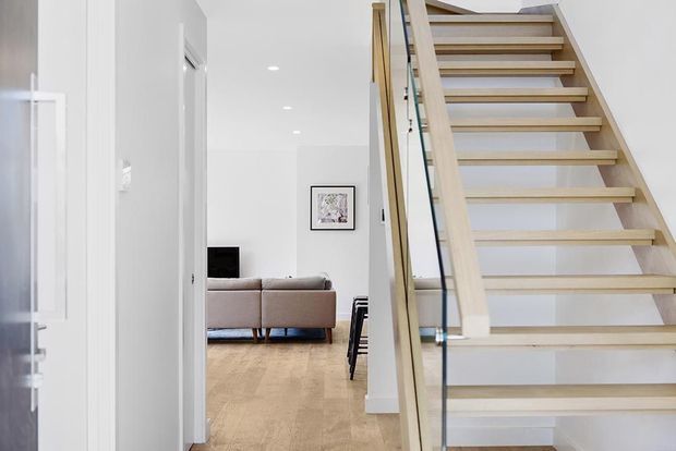 Bright minimalist residential interior showing an open-riser timber staircase with clear glass balustrade in the foreground, leading beside a white hallway toward a living room with a neutral sofa and timber flooring.