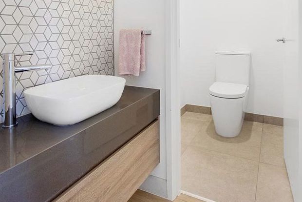 Bright contemporary bathroom featuring a floating timber-veneer vanity with a dark stone benchtop, white vessel basin, chrome tapware, geometric hexagonal wall tiles, and a separate toilet area with large beige floor tiles. A soft pink towel adds a subtle accent.