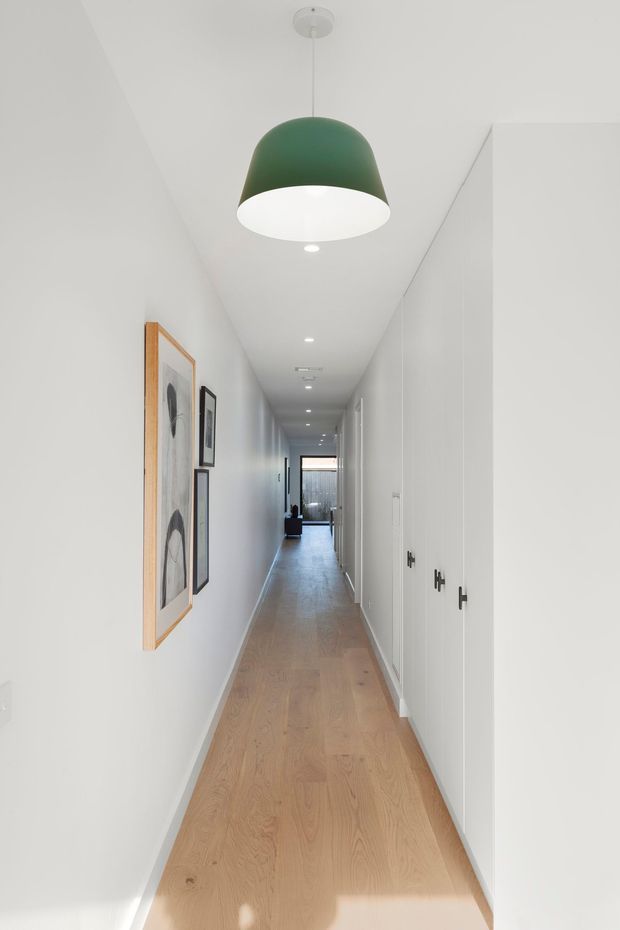 A long, narrow residential hallway with white walls and ceiling, pale timber flooring, recessed downlights, and a large green pendant light near the foreground. Full-height white built-in cabinetry with dark handles lines the right side, while framed artwork hangs on the left wall. The corridor leads toward a glazed opening and a darker living area at the far end.