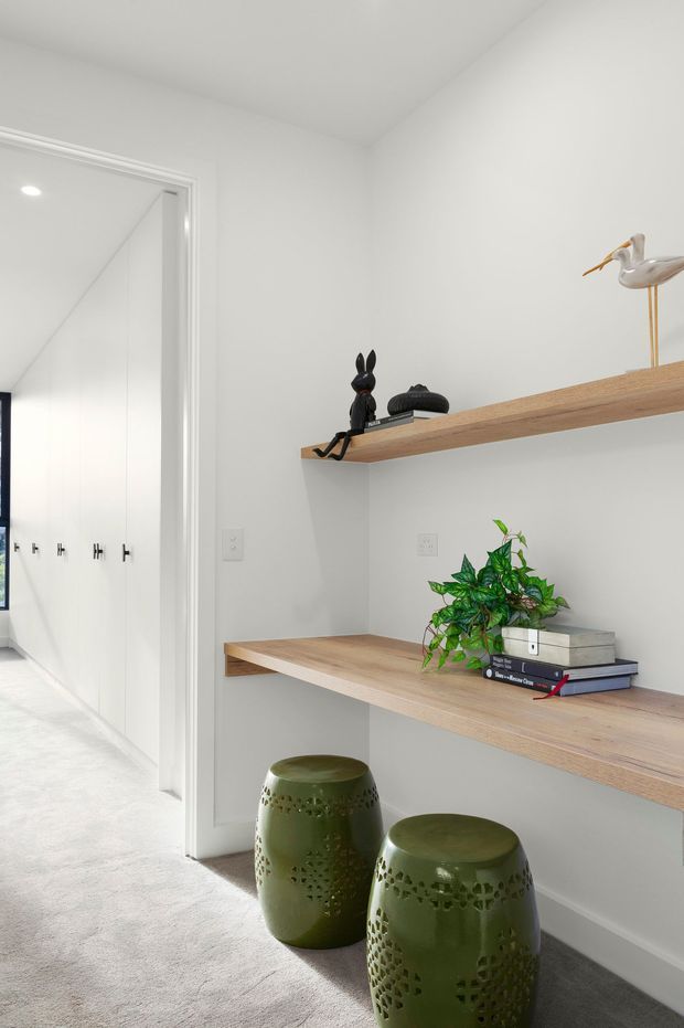 Bright minimalist hallway or landing with white walls, carpeted flooring, and built-in open timber shelving. The shelves display books, small sculptures, and a plant, with two green decorative stools positioned beneath. A white built-in wardrobe or storage run is visible through the doorway.