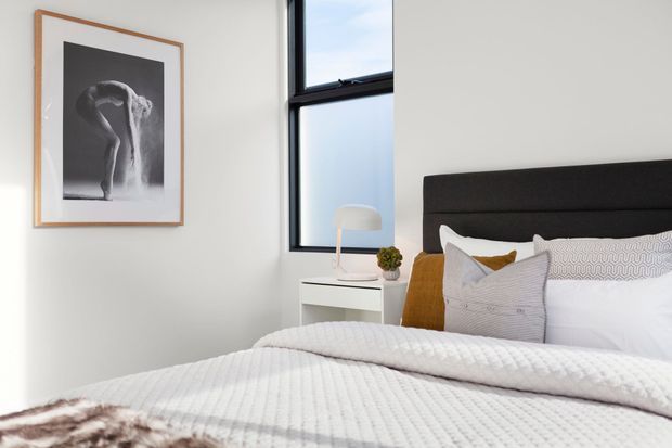 Bright contemporary bedroom with a neatly made bed, dark upholstered headboard, white quilted bedding, grey and mustard cushions, a white bedside table with lamp and plant, black-framed window, and framed monochrome artwork.