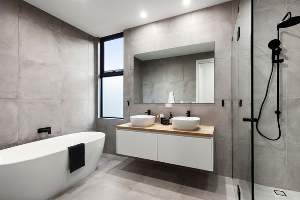 Contemporary bathroom with large-format grey stone-look wall and floor tiles, a freestanding white bathtub, floating white vanity with a timber benchtop and two vessel basins, frameless glass shower, black tapware and hardware, and a large recessed mirror.