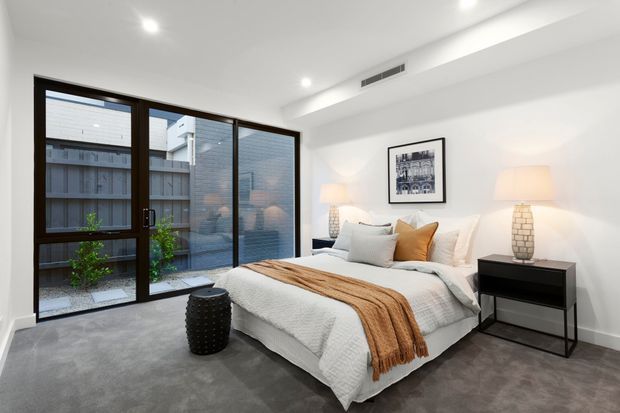 Bright contemporary bedroom with a white upholstered bed, layered white bedding, a rust-toned throw and cushions, dark bedside furniture, table lamps, recessed ceiling lighting, and a full-height dark-framed glazed door and window opening to a small courtyard.