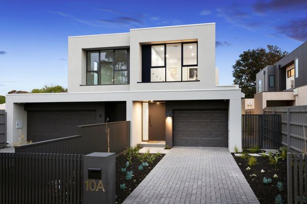 Street-facing exterior photograph of a contemporary two-storey townhouse with a clean white rendered facade, flat roofline, large black-framed windows, integrated double garage, covered entry, dark fencing, and a paved driveway. The neighbouring townhouse and landscaped front garden are also visible.