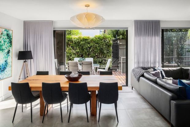 Bright contemporary open-plan dining and living area with a large timber dining table, black upholstered dining chairs, a dark sectional sofa, floor-to-ceiling sliding glazing, sheer curtains, and a covered outdoor courtyard visible beyond. A sculptural pendant light and large-format pale floor tiles complete the refined interior.