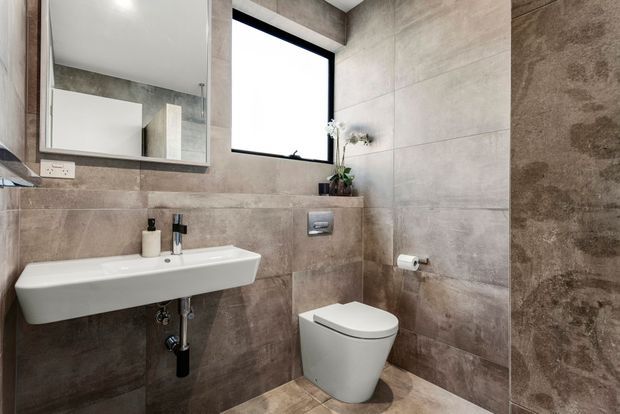 Compact contemporary bathroom with a wall-mounted white basin and toilet, large-format taupe and grey stone-look tiles on the walls and floor, a black-framed window, recessed toilet cistern, and a rectangular wall mirror. The space has a clean, refined, neutral finish with black metal accents and a small plant.