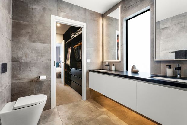Contemporary bathroom with large-format grey stone-look wall and floor tiles, a wall-hung white toilet, and a long floating vanity with matte white cabinetry and a dark benchtop. A large mirrored cabinet, black-framed window, recessed shelving, and a walk-in wardrobe visible through the doorway add functional storage and depth. Warm under-vanity lighting contrasts with the cool grey finishes.