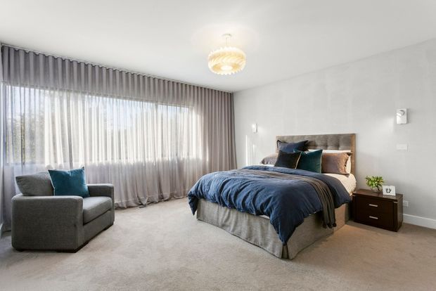 A spacious, softly lit contemporary bedroom with a large upholstered bed, blue and grey bedding, matching grey armchair, full-width sheer curtains, carpeted floor, bedside tables, and a sculptural pendant light. The neutral palette and layered textiles create a refined, comfortable atmosphere.