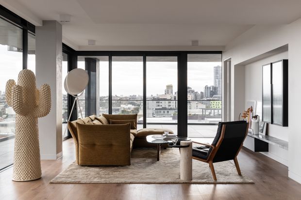 A refined high-rise living room with floor-to-ceiling black-framed glazing overlooking a dense city skyline. The room features warm timber flooring, a neutral textured rug, sculptural tan lounge chairs, a dark leather-and-wood accent chair, a low coffee table, a cylindrical stone pedestal, and minimalist wall-mounted cabinetry. A large woven cactus sculpture and a tripod floor lamp add distinctive decorative elements.