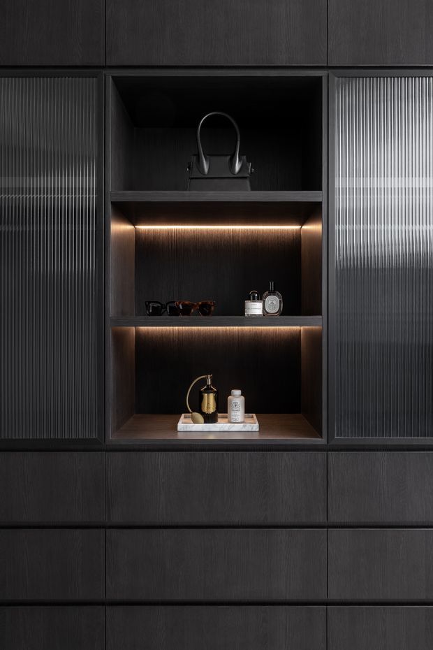 Close-up of a dark, sophisticated built-in storage wall with vertically grained black-stained timber cabinetry. The central open shelving niche is illuminated with warm integrated LED strip lighting and styled with a handbag, sunglasses, perfume bottles, and a small tray. Fluted translucent glass-fronted cabinet doors flank the shelving, creating a refined, moody feature joinery installation.