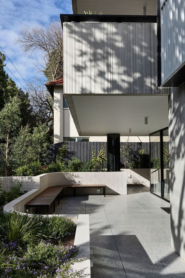 A contemporary multi-level residential courtyard and terrace viewed from outdoors. The space features large grey tiled paving, white brick planter and retaining walls, a built-in timber bench, landscaped garden beds, dark timber fencing, extensive glazing, and a projecting upper volume clad in vertically ribbed light-grey metal. Strong sunlight creates tree shadows across the facade and terrace.