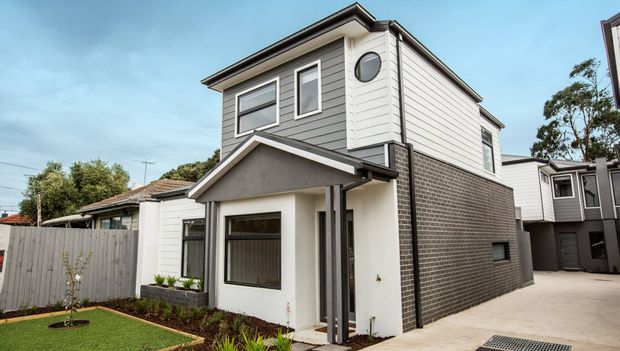 Exterior view of a contemporary two-storey attached townhouse or compact residential dwelling. The facade combines light grey horizontal weatherboard cladding, dark grey brickwork, white rendered walls, black-framed windows, and a small covered entry porch. A narrow landscaped front garden and concrete driveway are visible.