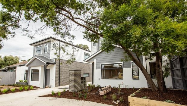 Exterior view of a contemporary multi-level residence with light grey weatherboard cladding, dark grey brickwork, contrasting trim, and a compact landscaped front garden. The home has a mix of pitched and low-slope roof forms, vertically arranged windows, a covered entry, and a pale concrete pathway, partially framed by mature trees.