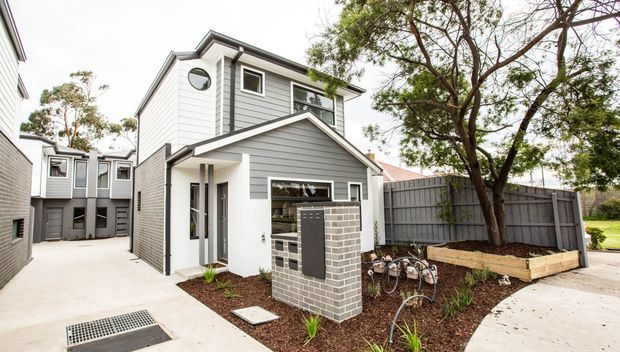 Exterior view of a compact multi-level contemporary townhouse with grey horizontal cladding, white trim, a pitched gable roof, and a small front entry porch. The property is arranged along a paved shared access path, with a low brick mailbox structure, timber-edged garden beds, bicycle parking, fencing, and mature trees.