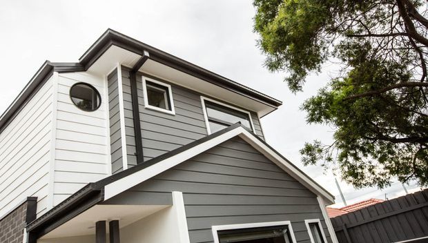 Exterior view of a contemporary multi-level home with overlapping pitched roof forms, dark grey horizontal weatherboard cladding, white trim, black-framed windows, and a covered entry canopy. Mature landscaping and a dark boundary fence are visible around the house.