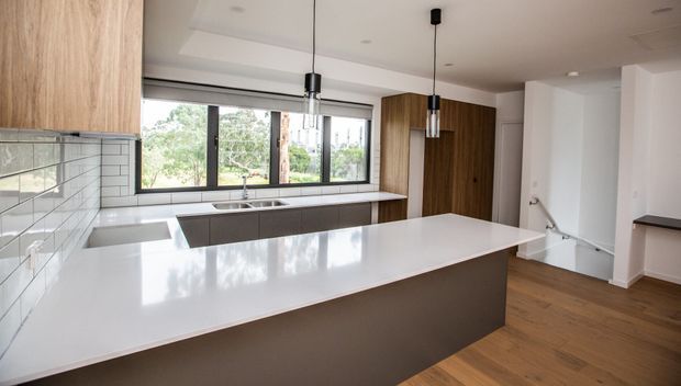 Modern open-plan kitchen with a large white island, dark integrated cabinetry, warm timber flooring and upper joinery, white subway-tile splashback, stainless-steel sink, and black-framed windows. Pendant lights hang above the island, with a bright, clean outlook to greenery beyond the glazing.