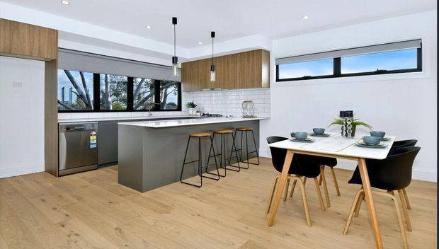 Bright open-plan kitchen and dining area with light timber flooring, white walls, black-framed windows, white subway-tile splashback, timber overhead cabinetry, and a central island with dark stools. A modern dining table and chairs sit beside the kitchen, with leafy trees visible through the glazing.