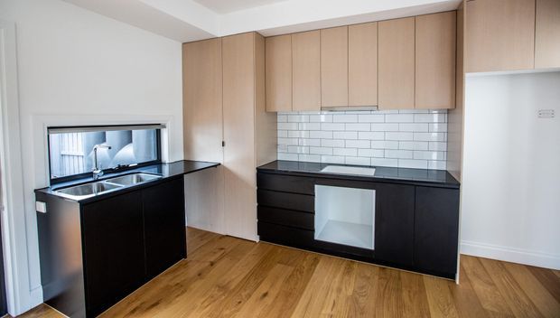 Compact contemporary kitchen with black lower cabinetry and benchtops, pale timber overhead cabinetry, white subway-tile splashback, timber flooring, stainless-steel sink, and integrated appliances. The room is bright and functional with a clean, modern fit-out.