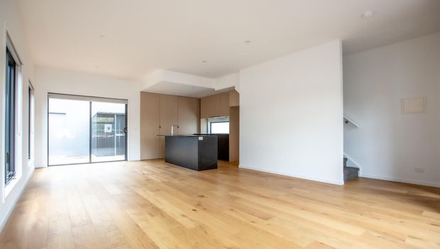 Bright open-plan interior with pale timber flooring, white walls, large glazed openings, and a compact contemporary kitchen featuring light wood cabinetry and a dark island. A partially concealed stairway is visible behind the dividing wall.