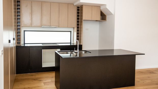 Contemporary kitchen with a large dark island, integrated sink and tap, dark lower cabinetry, pale timber overhead cabinetry, open wine-storage niches, and timber flooring. A wide horizontal window provides natural light, while a white stair or bulkhead structure is visible at right.